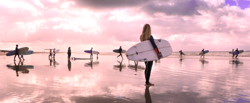 una surfista in riva al mare e sullo sfondo altri surfisti. L'immagine è in tonalità rosa. 