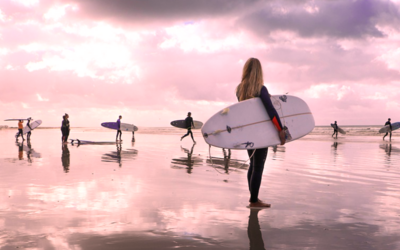 una surfista in riva al mare e sullo sfondo altri surfisti. L'immagine è in tonalità rosa. 