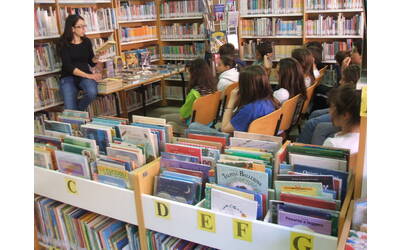 all'interno di una biblioteca una ragazza seduta su un tavolino legge un libro a un gruppo di bambine e bambini seduti
