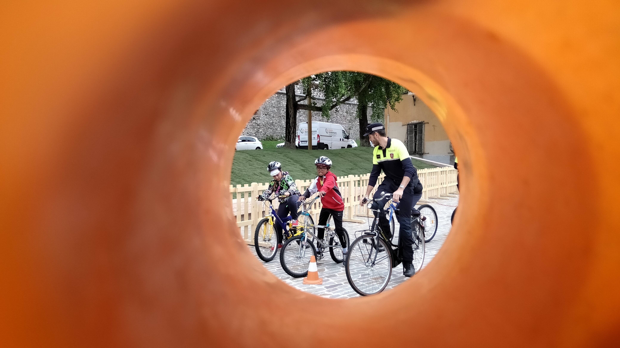 un agente di polizia locale in bicicletta affianca un bambino e una bambina in bicicletta lungo un percorso