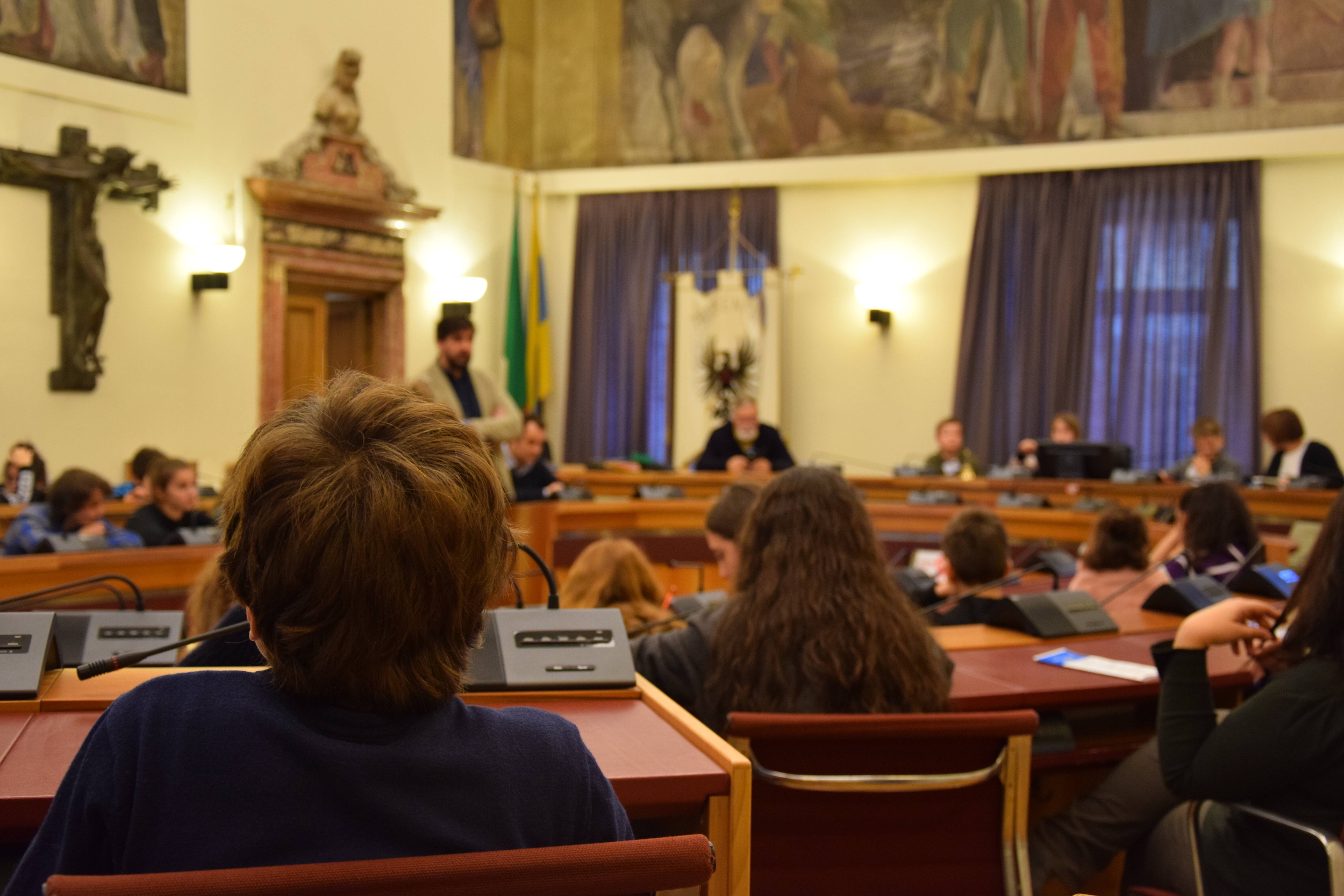 ragazzi e ragazze seduti all'interno dell'aula del consiglio comunale ascoltano un adulto che parla
