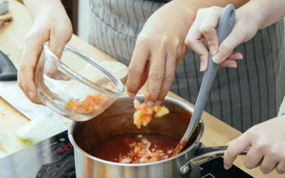 Le mani di una donna tengono la padella e mescolano il sugo all'interno, mentre quelle di un'altra ci versano del cibo all'interno.