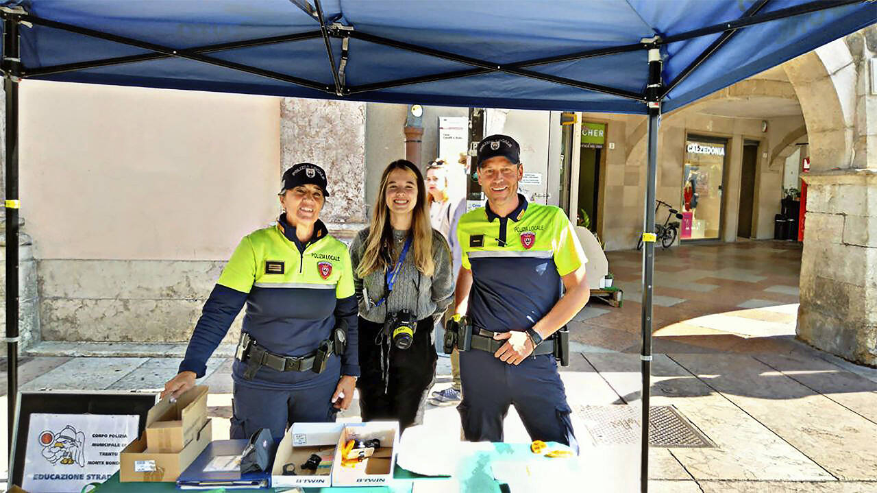una ragazza con al collo una macchina fotografica e due agenti della polizia locale sorridono al fotografo presso lo stand della Polizia locale