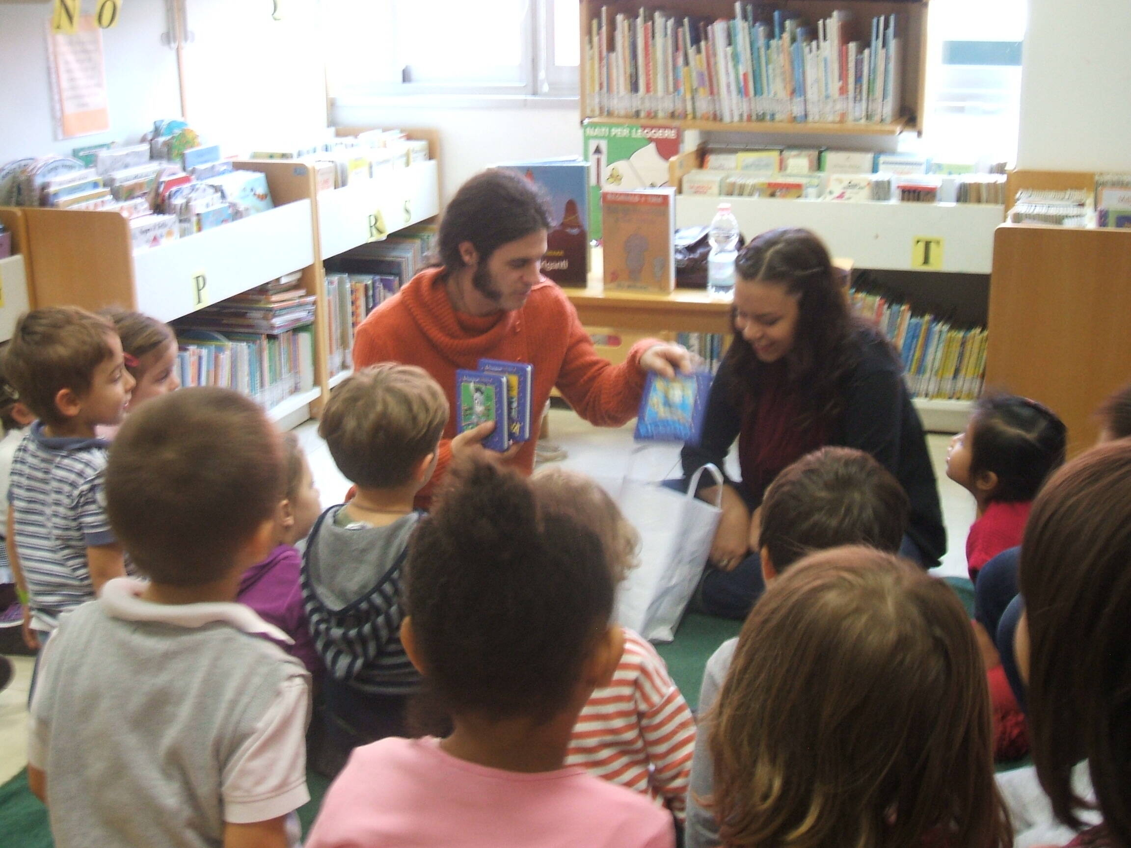 un gruppetto di bambini seduti per terra osservano il bibliotecario e la maestra che mettono in una borsa alcuni libretti