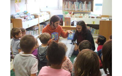un gruppetto di bambini seduti per terra osservano il bibliotecario e la maestra che mettono in una borsa alcuni libretti