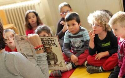una maestra di spalle, mostra un libro illustrato a sette bambini e bambine seduti per terra