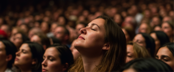 varie persone in un teatro, una donna in primo piano con il viso rivolto verso l'alto sembra in ascolto